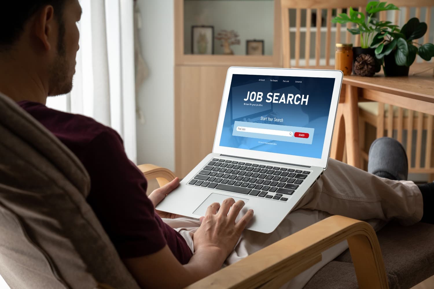 A man sitting in a chair at home using a laptop for online job searching, with a job search webpage displayed on the screen.